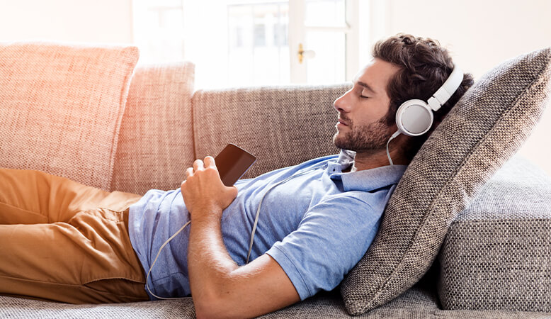 Side view of man laying on brown couch listening to music with white earphones